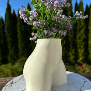 May include: White ceramic vase shaped like a human body, with a bouquet of purple flowers.