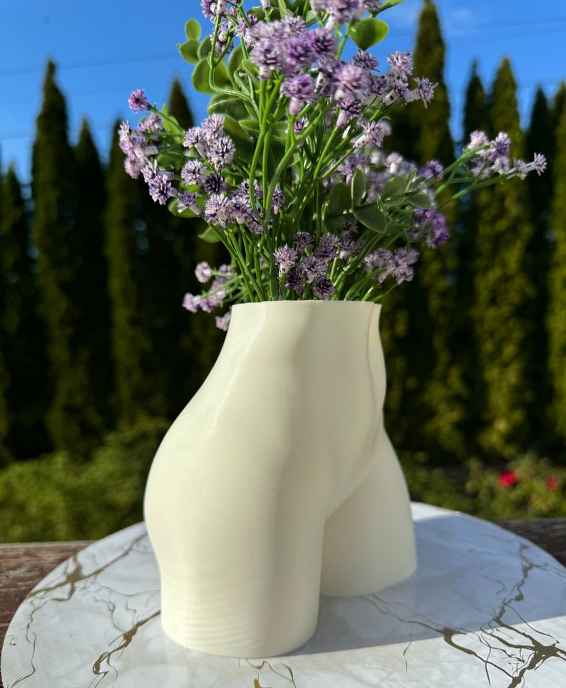 May include: White ceramic vase shaped like a human body, with a bouquet of purple flowers.