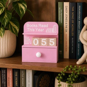 May include: A pink wooden book counter with the text "Books Read This Year" and the number "055" displayed on wooden blocks. The counter has a drawer with a white knob. Books are visible in the background.