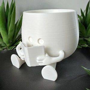 May include: White ceramic planter shaped like a person sitting and reading a book. The planter has a round body and two legs.