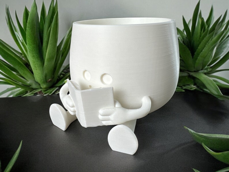 May include: White ceramic planter shaped like a person sitting and reading a book. The planter has a round body and two legs.