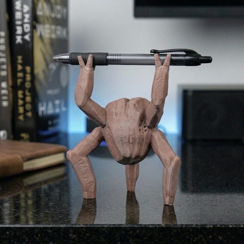 May include: A brown, 3D-printed pen holder shaped like a creature with four legs and arms holding a pen. The pen is black and silver. The creature has a textured surface and the word "HAIL" is printed on one of its legs.