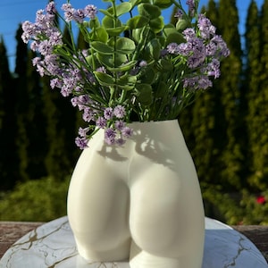 May include: White ceramic vase shaped like a human bottom, with a bouquet of purple flowers.