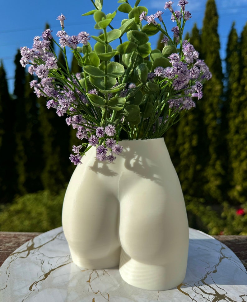 May include: White ceramic vase shaped like a human bottom, with a bouquet of purple flowers.