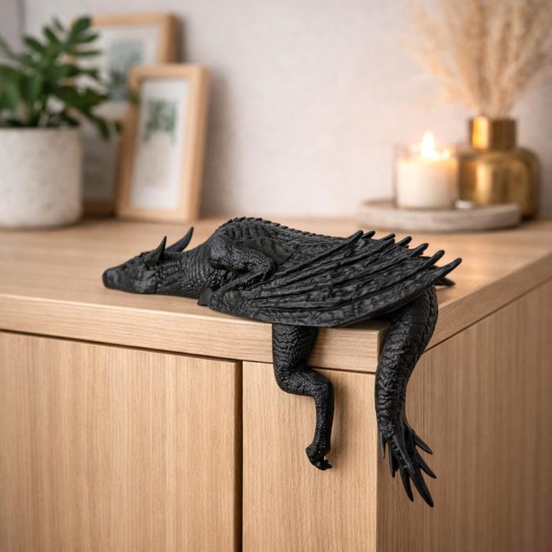 May include: A black dragon figurine with detailed scales, wings, and claws, resting on a wooden surface. The dragon's body is positioned to hang over the edge, creating a decorative accent. The figurine is a fantasy-themed home decor item.