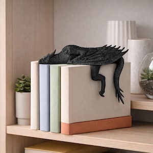 May include: A black dragon bookend rests atop a stack of books. The dragon has detailed wings and a long tail that hangs down the side. The books are various colors, including beige, blue, and green. The bookend adds a fantasy theme to any bookshelf.