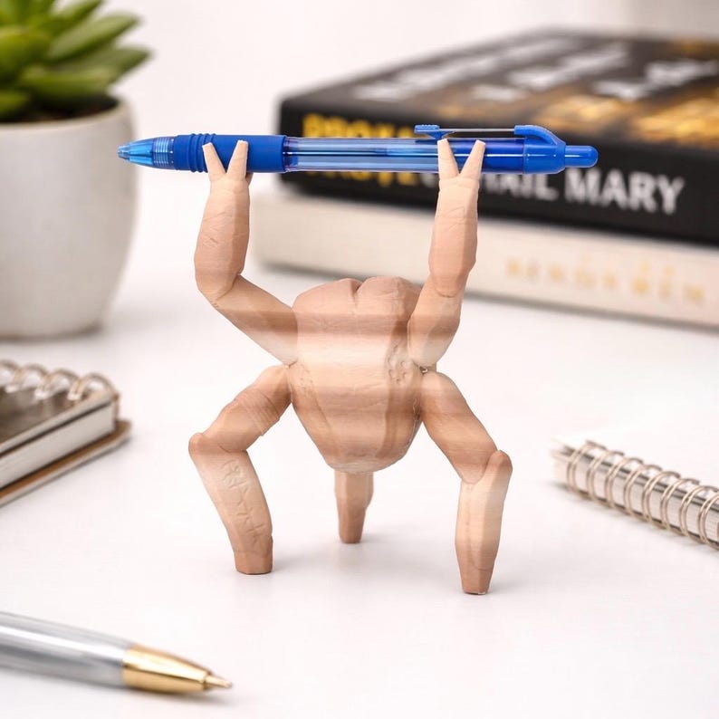 May include: A novelty pen holder shaped like a wooden figure with arms raised, holding a blue pen. The figure has four legs and is positioned on a white surface. A succulent plant in a white pot and a spiral notebook are in the background.