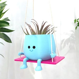 May include: A light blue hanging planter shaped like a smiling face with legs and arms. The planter is holding a green plant and is suspended from a white rope. The planter is sitting on a pink platform.