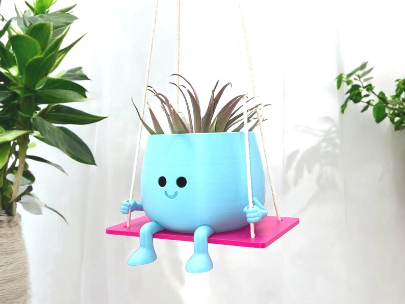 May include: A light blue hanging planter shaped like a smiling face with legs and arms. The planter is holding a green plant and is suspended from a white rope. The planter is sitting on a pink platform.