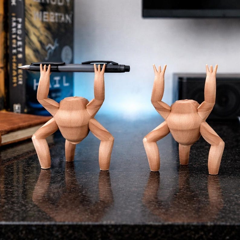 May include: Two wooden pen holders shaped like figures, one holding a pen aloft. The figures are light brown with four legs and arms raised. The pen is black and gray. The figures are on a dark, reflective surface.
