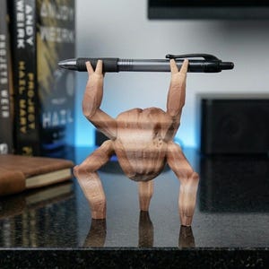 May include: A brown, 3D-printed pen holder shaped like a figure lifting a pen overhead. The pen is black and silver. The figure has a textured, wood-like appearance and is standing on a dark surface.
