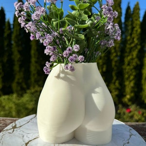 May include: White ceramic vase shaped like a human body, with a bouquet of purple flowers.