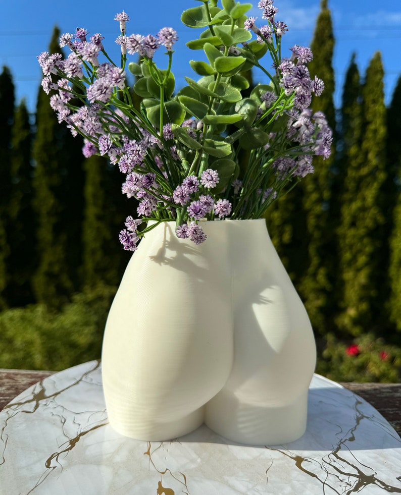May include: White ceramic vase shaped like a human body, with a bouquet of purple flowers.
