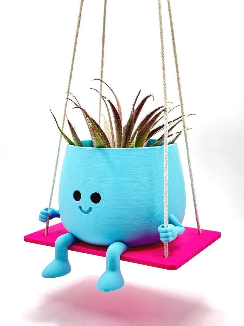 May include: A light blue hanging planter shaped like a smiling face with arms and legs, holding onto a pink swing. The planter has a small air plant inside.