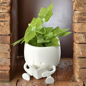 May include: White ceramic planter shaped like a person sitting and reading a book. The planter has a smiling face and is holding a white book. The planter is filled with green leafy plants.