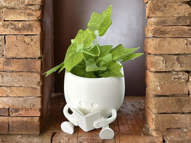 May include: White ceramic planter shaped like a person sitting and reading a book. The planter has a smiling face and is holding a white book. The planter is filled with green leafy plants.
