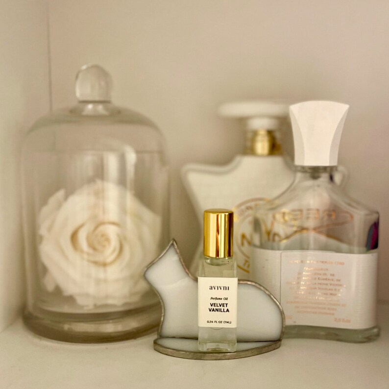 May include: A perfume oil bottle with a gold cap and the text "avivni Velvet Vanilla" sits on a cat-shaped holder. A glass cloche with a white rose and other perfume bottles are in the background.