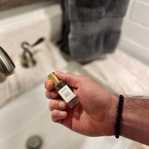 May include: A small perfume bottle with a gold cap is held in a hand. The bottle has a white label with black text that reads "avani Velvet Vanilla". A black beaded bracelet is on the wrist. The bottle contains 10 ml.