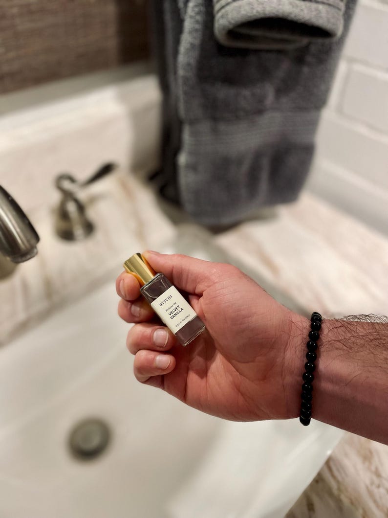May include: A small perfume bottle with a gold cap is held in a hand. The bottle has a white label with black text that reads "avani Velvet Vanilla". A black beaded bracelet is on the wrist. The bottle contains 10 ml.