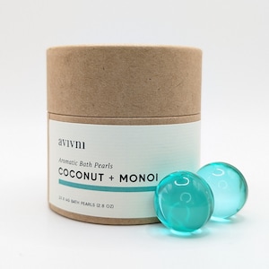 May include: A brown cardboard tube with a white label that reads "avivni Aromatic Bath Pearls COCONUT + MONOI 15 X 4G BATH PEARLS (2.8 OZ)". Two teal bath pearls are in front of the tube.