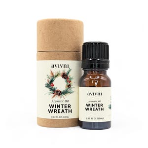 May include: A brown cardboard tube with a white label that says "avivnı Aromatic Oil WINTER WREATH 0.33 FL OZ (10ML)". The label has a watercolour illustration of a winter wreath with pine branches, pine cones, and red berries. A small brown glass bottle with a black cap and a white label that says "avivnı Aromatic Oil WINTER WREATH 0.33 FL OZ (10ML)" is in front of the tube.