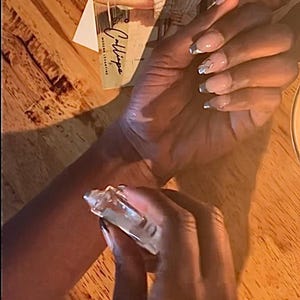 May include: A clear perfume bottle being sprayed onto a wrist. The nails are painted with a light pink and silver design. A card with the word "Calliope" is visible in the background.