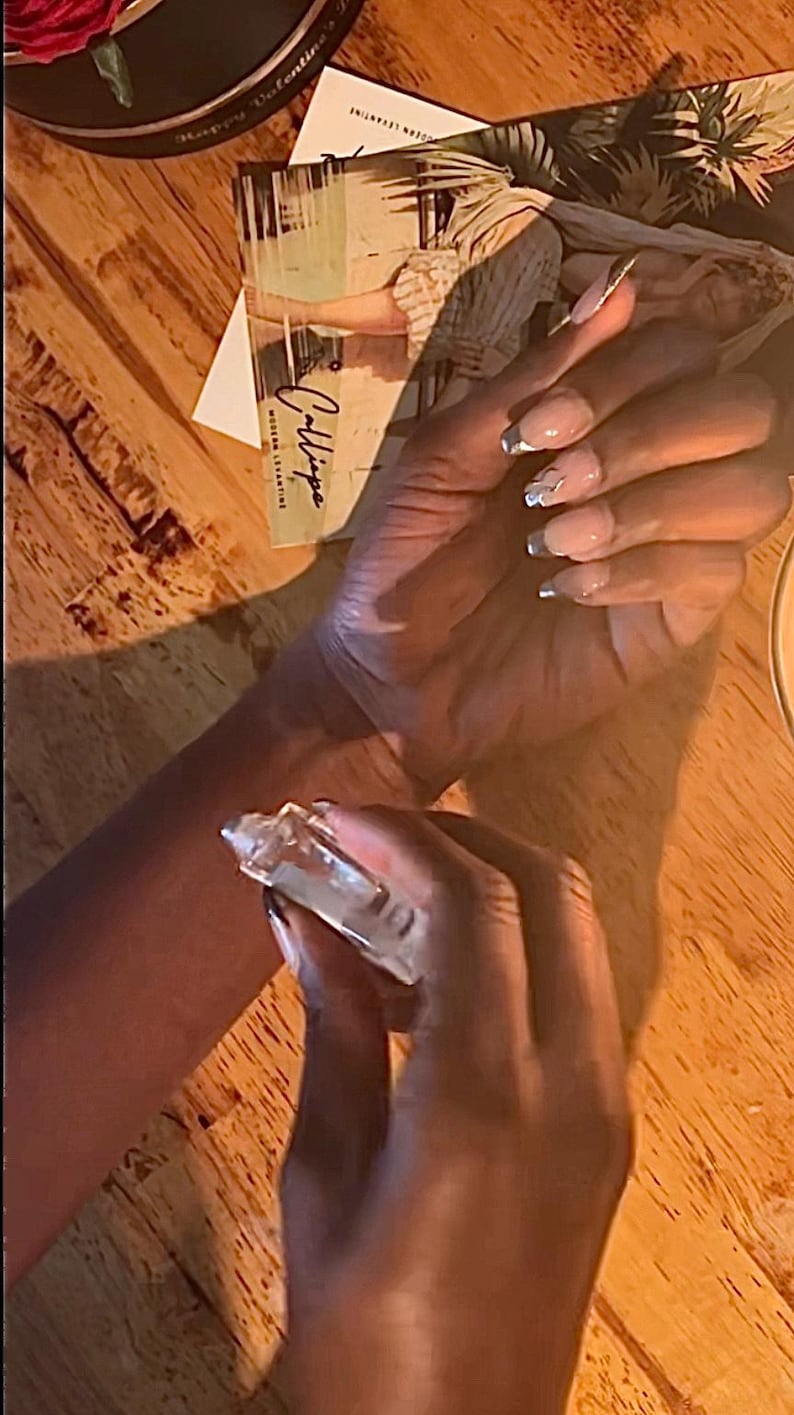 May include: A clear perfume bottle being sprayed onto a wrist. The nails are painted with a light pink and silver design. A card with the word "Calliope" is visible in the background.