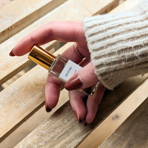 May include: A small, clear perfume bottle with a gold cap is held in a hand with dark red nail polish. The bottle has a white label with black text. The background is a light-coloured wooden surface.