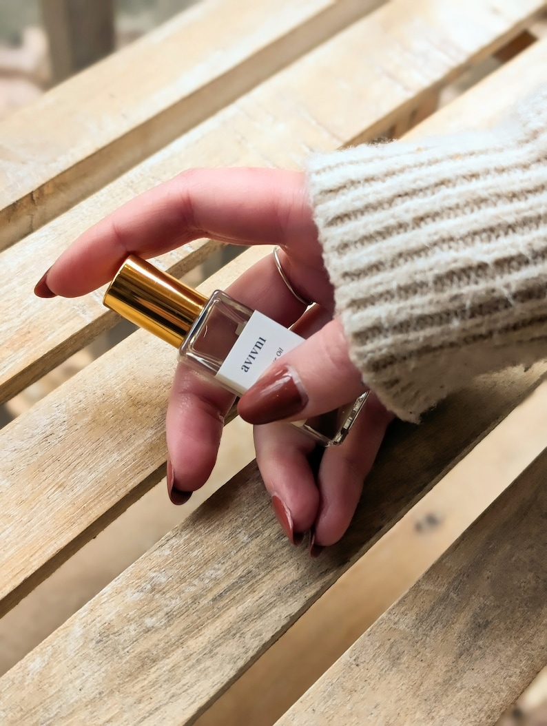 May include: A small, clear perfume bottle with a gold cap is held in a hand with dark red nail polish. The bottle has a white label with black text. The background is a light-coloured wooden surface.