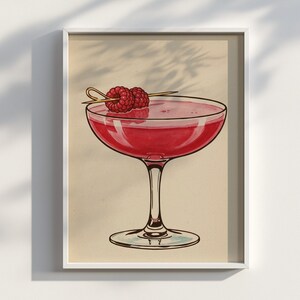 May include: A framed illustration of a cocktail in a coupe glass. The drink is a vibrant red color, garnished with two raspberries on a wooden skewer. The artwork is set against a neutral background.