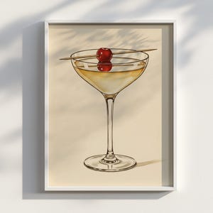 Op de afbeelding: Een ingelijste print met een cocktail in een glas met steel, gegarneerd met een rode kers op een houten spies. De drank heeft een lichtgele kleur en staat tegen een crèmekleurige achtergrond.