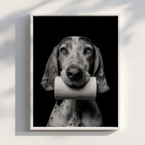 Op de afbeelding: Zwart-wit ingelijste print met een hond die een rol toiletpapier in zijn bek houdt. De hond heeft een gevlekte vacht en lange oren. De achtergrond is effen zwart.