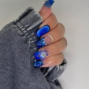 Puede incluir: Primer plano de una mano con arte de u&ntilde;as azul y blanco. Las u&ntilde;as son largas y de forma cuadrada. El arte de u&ntilde;as presenta una variedad de dise&ntilde;os, que incluyen estampado de leopardo, flores y brillo.