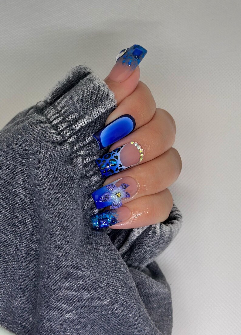 Puede incluir: Primer plano de una mano con arte de u&ntilde;as azul y blanco. Las u&ntilde;as son largas y de forma cuadrada. El arte de u&ntilde;as presenta una variedad de dise&ntilde;os, que incluyen estampado de leopardo, flores y brillo.