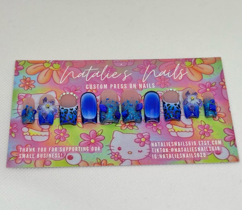 Puede incluir: U&ntilde;as postizas azules y blancas con dise&ntilde;o de estampado de leopardo, purpurina y pedrer&iacute;a. Las u&ntilde;as se muestran en una tarjeta con el texto "Natalie's Nails Custom Press On Nails" y la informaci&oacute;n de contacto.