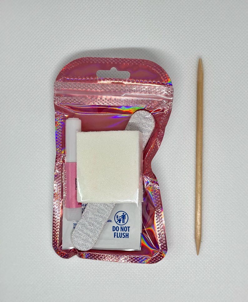 Puede incluir: Una bolsa hologr&aacute;fica rosa que contiene suministros de arte de u&ntilde;as, incluyendo una lima de u&ntilde;as, una peque&ntilde;a botella de pegamento, un buffer de u&ntilde;as blanco y un peque&ntilde;o paquete blanco con el texto "No tirar por el inodoro". Un empujador de cut&iacute;culas de madera est&aacute; al lado de la bolsa.