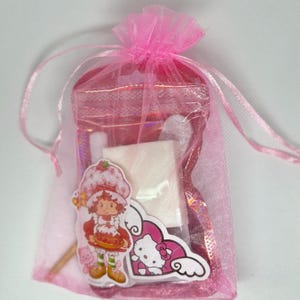 Puede incluir: Una bolsa de tul rosa llena de peque&ntilde;os regalos. La bolsa contiene una caja cuadrada blanca, una pegatina rosa de Hello Kitty, una pegatina de Strawberry Shortcake y un palo de madera.