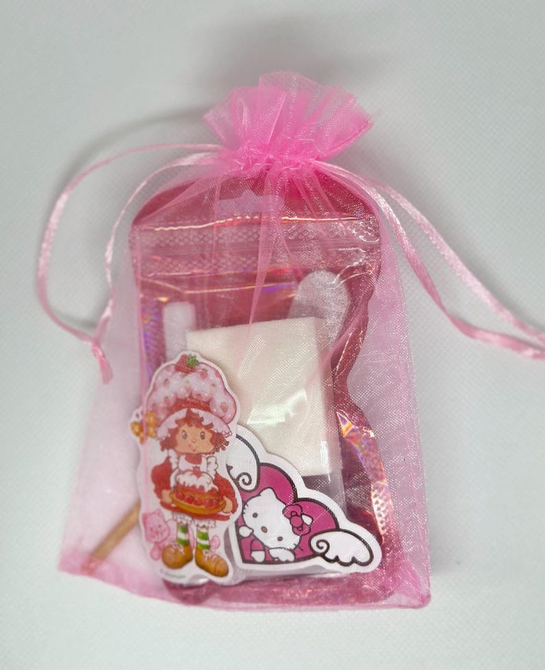 Puede incluir: Una bolsa de tul rosa llena de peque&ntilde;os regalos. La bolsa contiene una caja cuadrada blanca, una pegatina rosa de Hello Kitty, una pegatina de Strawberry Shortcake y un palo de madera.