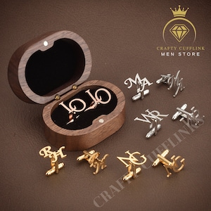 Può includere: Un set di sei gemelli personalizzati in tonalità oro e argento. I gemelli sono in una scatola di legno e presentano diverse iniziali. La scatola è aperta e i gemelli sono disposti intorno ad essa. Il testo "CRAFTY CUFFLINK MEN STORE" è visibile nell'angolo in alto a destra dell'immagine.