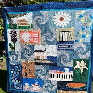 May include: A handmade quilt with a blue border and the name "DAISY PARKER" across the top. The quilt features various squares with images of a bench, a daisy, books, a rose, a clock, a house, a piano, and a lily.