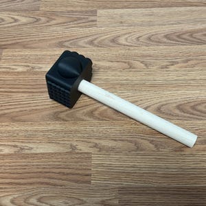 May include: A meat tenderizer mallet featuring a black, square-shaped head with a textured, grid-like surface on one side and a smooth, rounded top, attached to a light-colored, cylindrical wooden handle for a secure and comfortable grip.