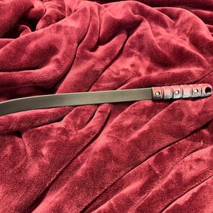 May include: A black, curved blade with a handle featuring a series of silver-colored rivets and a hole at the end. The blade is lying on a deep red, plush fabric.