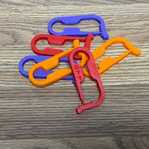 May include: A collection of five plastic bag clips in purple, red, and orange. Each clip features a long, narrow, oval-shaped opening with a serrated edge on one side for gripping. The red clip in the foreground has the letters DTT embossed on it.
