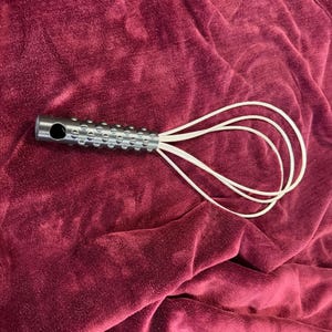 Pode incluir: Um batedor de metal com uma alça texturizada e cordas brancas. A alça é prateada com uma série de pequenas reentrâncias espaçadas uniformemente. As cordas são presas à alça e se estendem para fora.