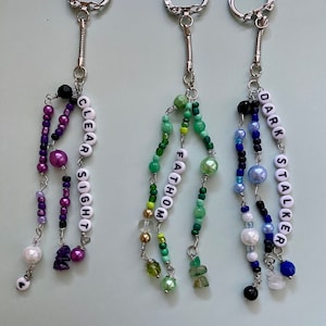 May include: Three beaded keychains with silver key rings. The keychains are made with different coloured beads and have the words "CLEAR SIGHT", "FATHOM", and "DARK STALKER" spelled out on them.