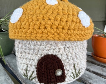 May include: A crocheted mushroom house with a brown door and white spots on the top. The house is made of beige and brown yarn.