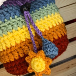May include: A rainbow crocheted drawstring bag with a white fluffy top, a purple drawstring, and a blue raindrop and yellow sun charm.