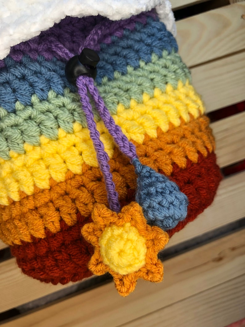 May include: A rainbow crocheted drawstring bag with a white fluffy top, a purple drawstring, and a blue raindrop and yellow sun charm.