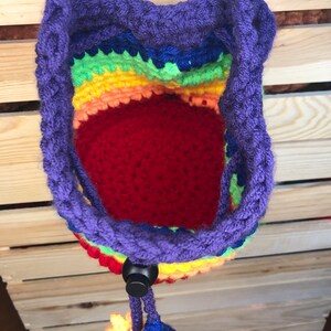 May include: A rainbow crocheted drawstring bag with a purple cord and a black toggle closure. The bag has a red lining and is hanging from a wooden surface.