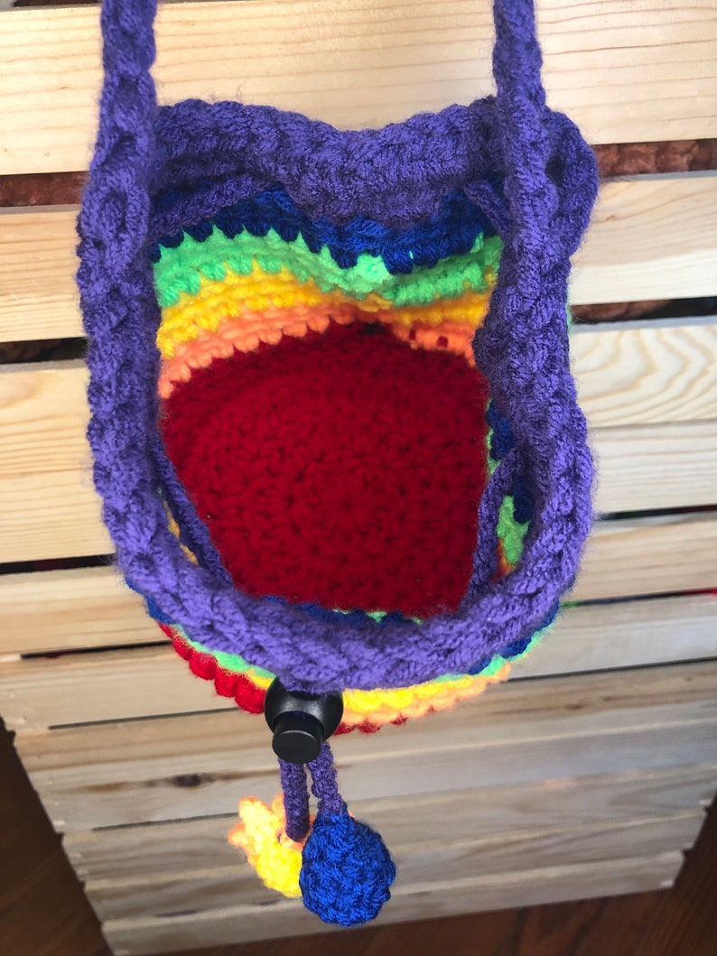 May include: A rainbow crocheted drawstring bag with a purple cord and a black toggle closure. The bag has a red lining and is hanging from a wooden surface.